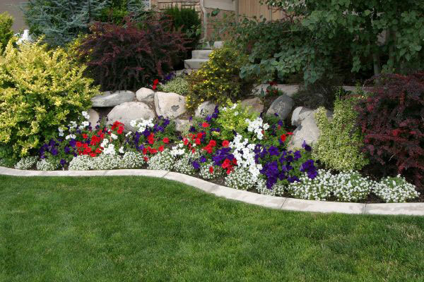 Colorful flower bed with red, white, and purple blooms, surrounded by greenery and bordered by a manicured lawn.