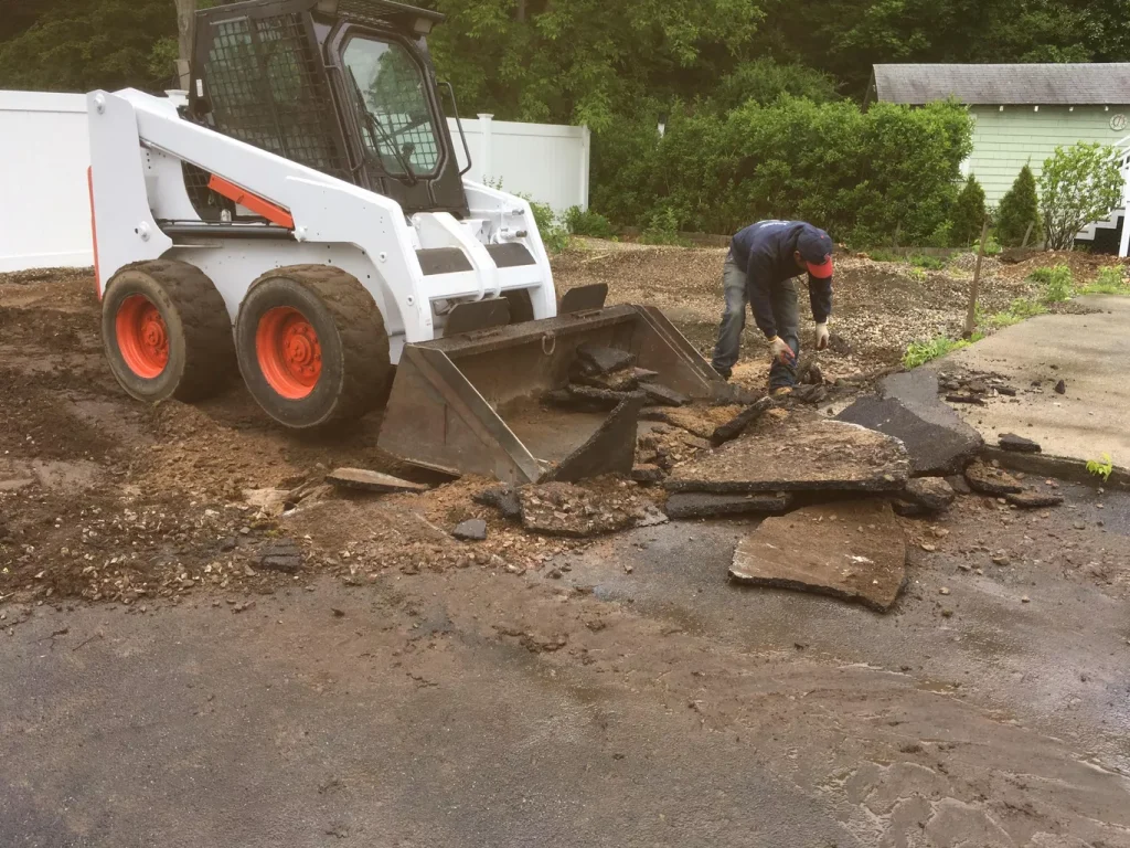 A person removes broken asphalt near a skid steer loader on a work site.