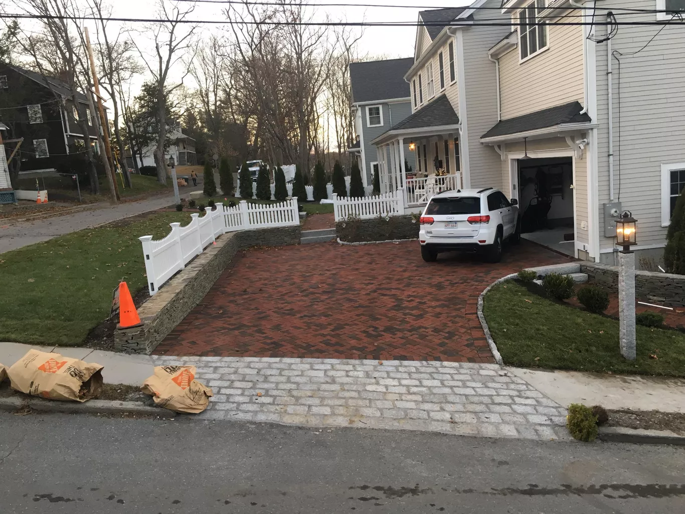 House with brick driveway, white SUV parked, white fence, and leaf bags on curb.