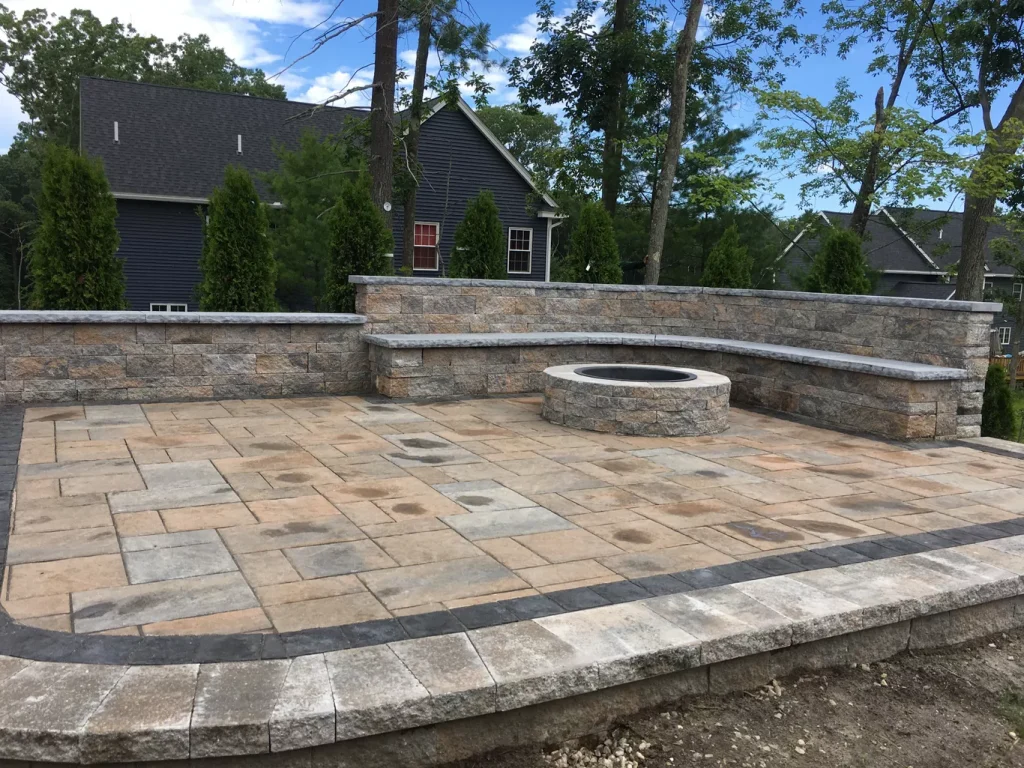 Stone patio with fire pit, curved bench, and surrounding trees near residential house.