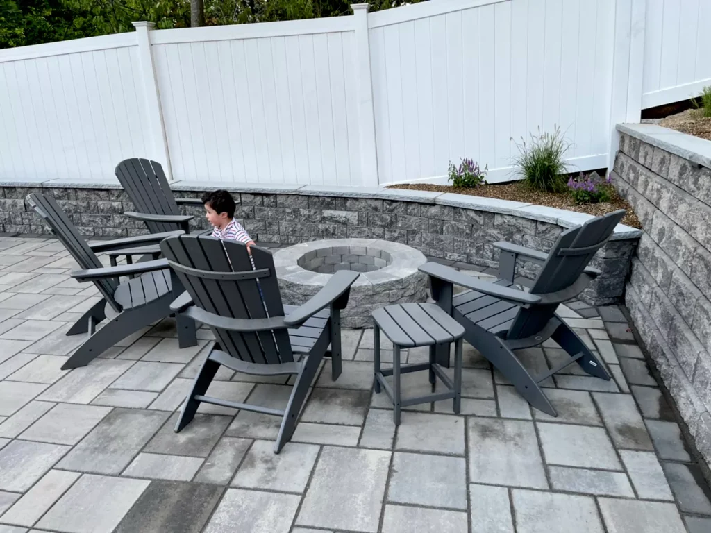 Patio with grey Adirondack chairs around a stone fire pit, a child sitting on one chair, and a white privacy fence.