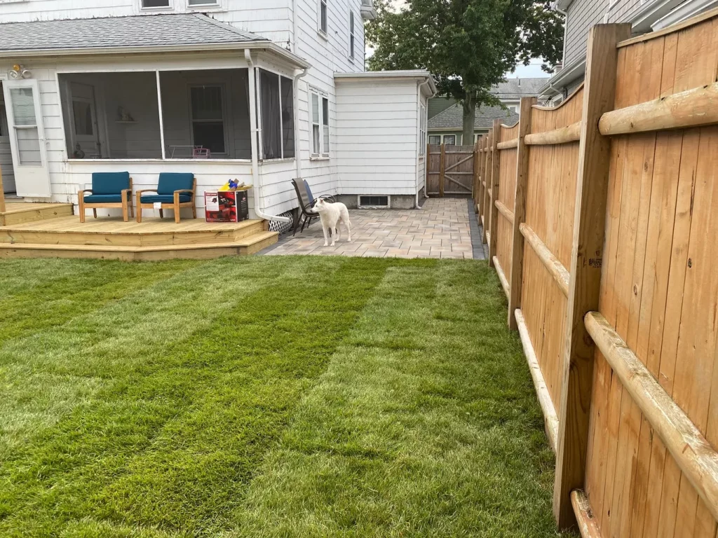 Backyard with green grass, wooden deck, patio area, two chairs, and a white dog standing near a wooden fence.