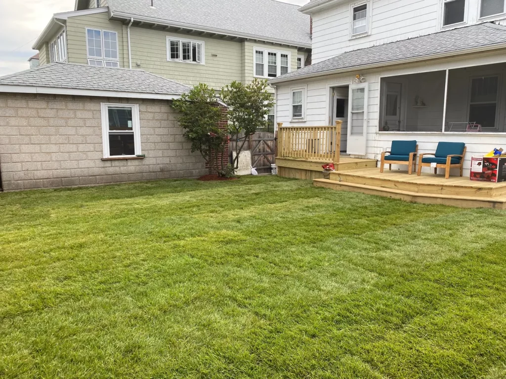 Backyard with green lawn, wooden deck, two blue chairs, and adjacent house.