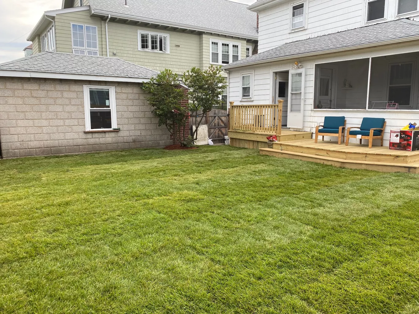 Backyard with green lawn, wooden deck, two blue chairs, and adjacent house.