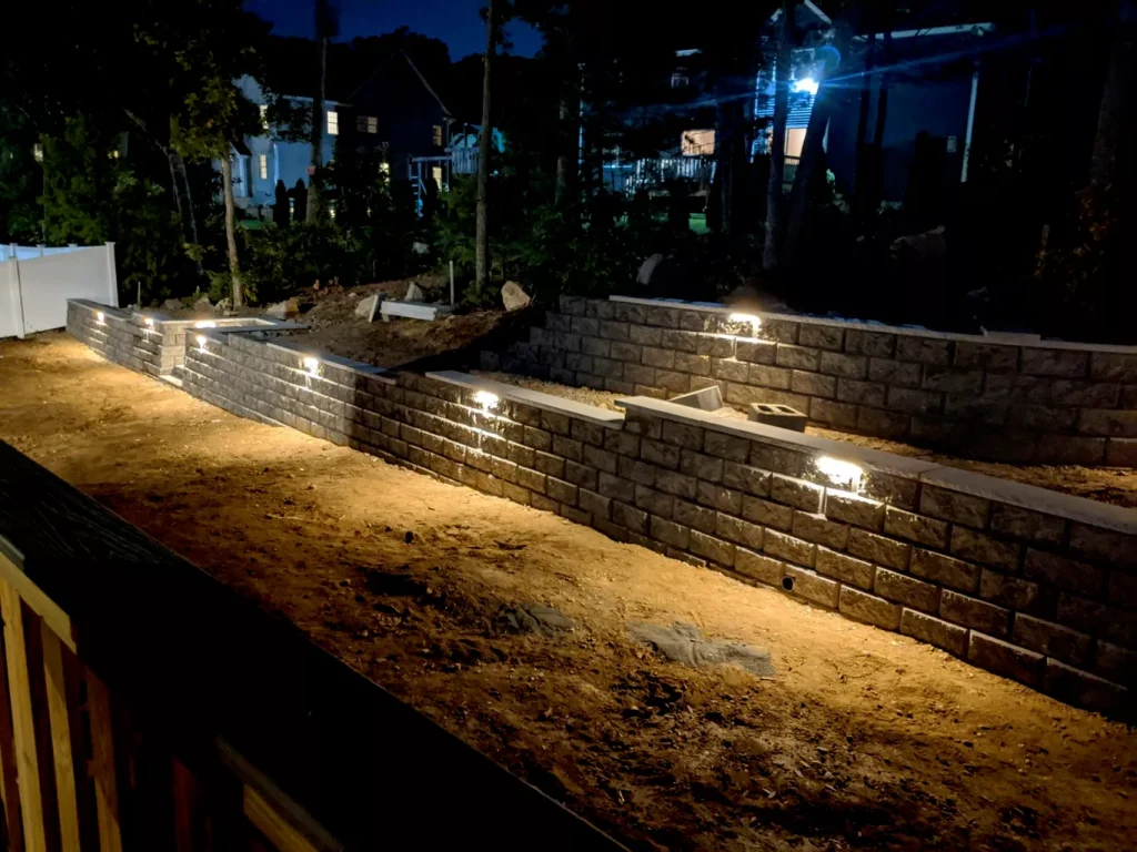 Retaining wall with embedded lights illuminating a dirt pathway, surrounded by trees and houses at night.