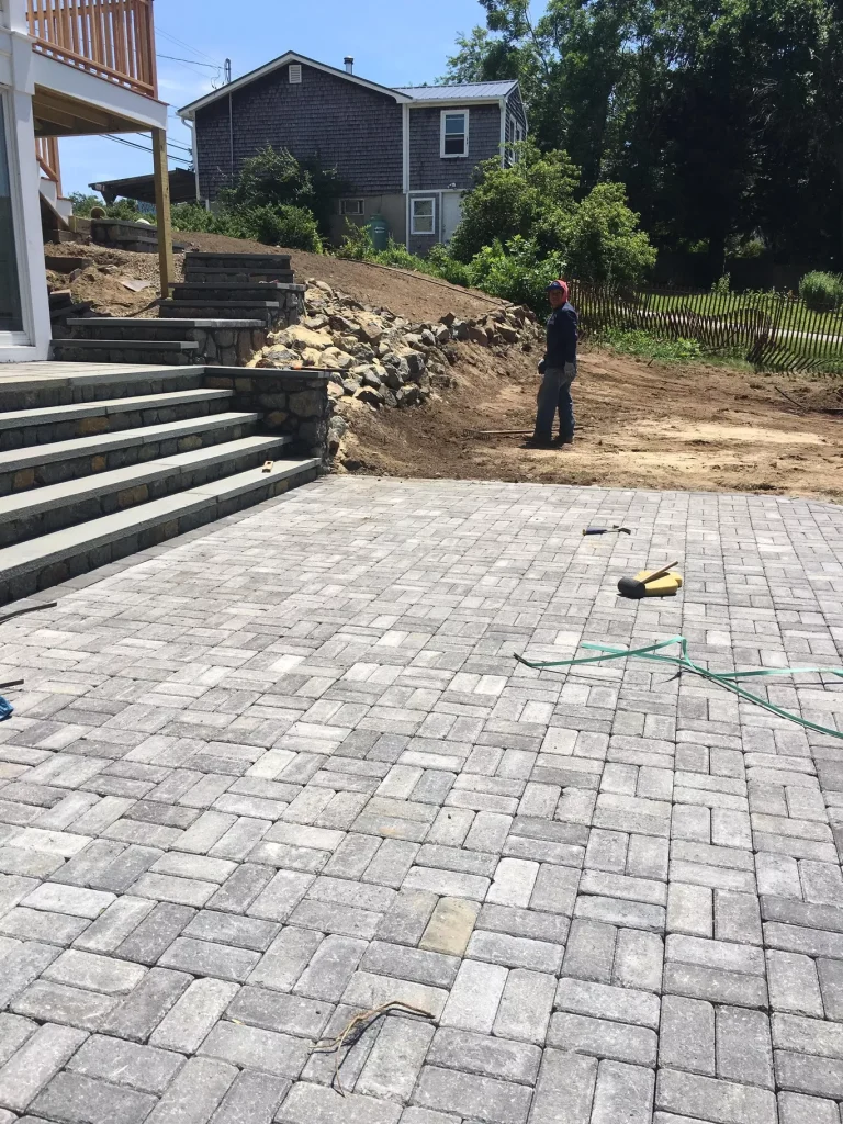 Newly laid stone patio with steps, a person standing nearby, and a house and trees in the background.