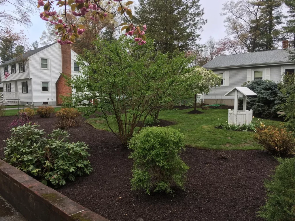 Well-maintained yard with a white wishing well, shrubs, and two white houses in the background.