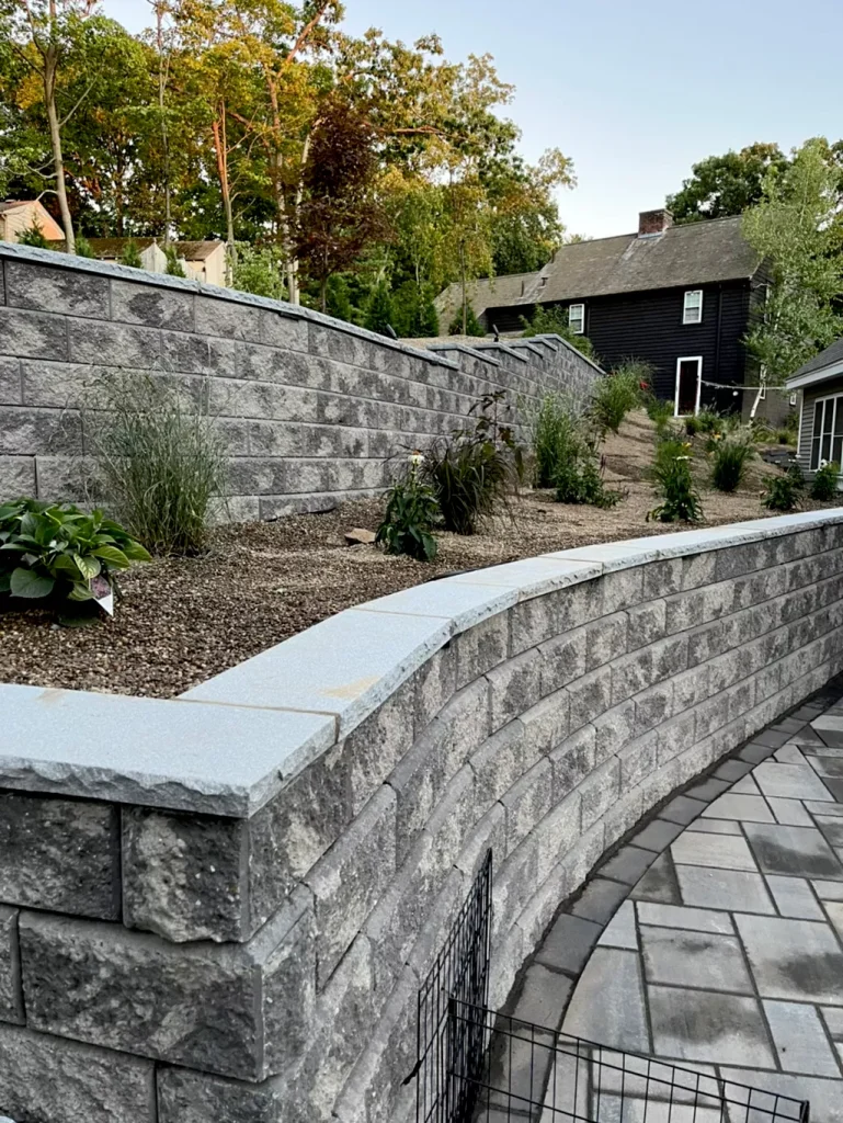 Stone retaining wall with landscaped plants, trees, and a dark house in the background.