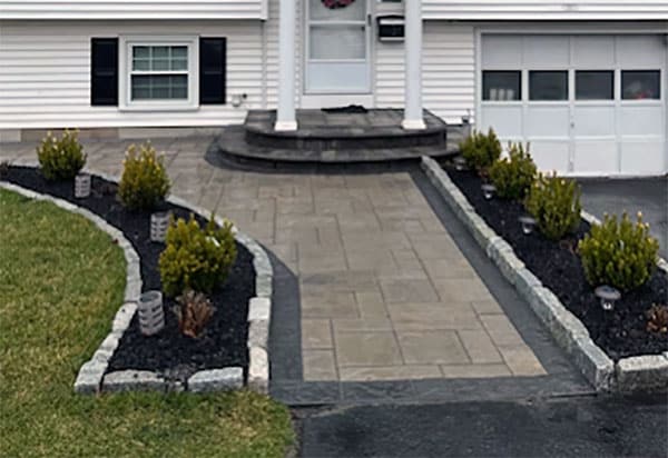 Front entrance with paved walkway, flanked by trimmed bushes and garden beds, leading to a white house with a garage.